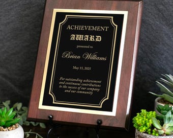 May include: A rectangular wooden award plaque with a dark brown finish and a black center plate. The plate features gold text reading ACHIEVEMENT AWARD, presented to Brian Williams, May 15, 2025, and a message regarding outstanding contributions.