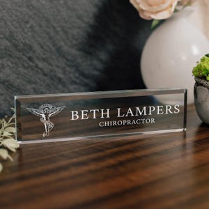 May include: A rectangular clear glass desk nameplate featuring the engraved white text BETH LAMPERS and CHIROPRACTOR. To the left of the name is a white engraved graphic of a winged figure with a banner reading HEALTH CHIROPRACTIC.