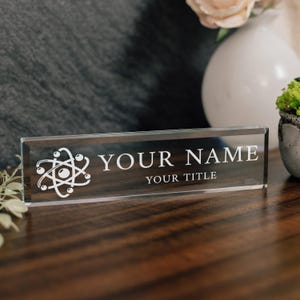 Può includere: Una targhetta da scrivania rettangolare in vetro trasparente con un simbolo atomico bianco inciso sul lato sinistro, recante il testo "YOUR NAME" in grandi lettere maiuscole e "YOUR TITLE" in maiuscole più piccole sul davanti.