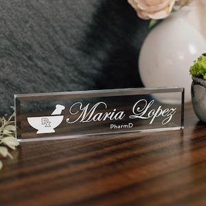 May include: A clear glass nameplate with the name "Maria Lopez" in elegant script. Below the name is "PharmD". A mortar and pestle symbol is on the left. The nameplate is rectangular and sits on a wooden surface.
