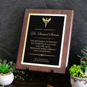 May include: A rectangular wooden plaque with a dark brown finish featuring a black center plate with gold lettering and a caduceus symbol. The text reads: In grateful appreciation to Dr. Samuel Stevens, followed by a message of thanks from the staff.