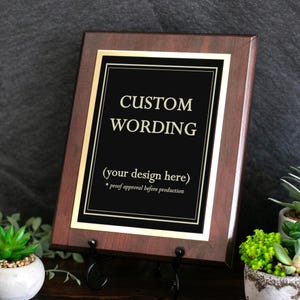 May include: A rectangular wooden plaque with a dark brown finish featuring a central black plate with gold trim. The plate displays the text CUSTOM WORDING, (your design here), and * proof approval before production in gold lettering.