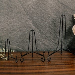 May include: Three black metal display stands of varying heights, each featuring a tripod design with curved feet and a central vertical support bar. These wire easels are constructed from thin, dark metal rods and are arranged in a row.