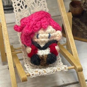 May include: A miniature crochet doll with bright pink hair, red and black clothing, and blue eyes, is seated in a small wooden beach chair with a cream-colored crocheted seat. The doll is the focal point of the image.