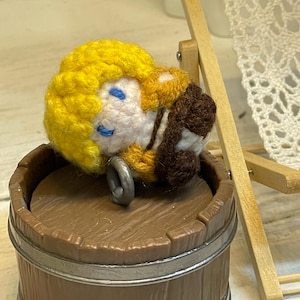 May include: A crocheted doll with yellow hair, blue eyes, and a brown and yellow outfit is lying on top of a wooden barrel. The doll is wearing a brown and yellow outfit and has a metal ring attached to its arm.