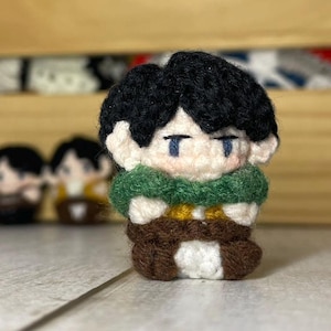 Op de afbeelding: Een gehaakt pluche speelgoed van een personage met zwart haar, een groene sjaal en een bruine broek. Het speelgoed heeft een wit gezicht met blauwe ogen en een kleine, gestikte glimlach.