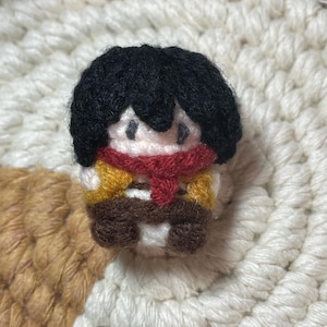 May include: A crocheted amigurumi figure of a person with black hair, a red scarf, and a yellow and brown outfit.