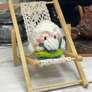 May include: A crocheted gnome doll with white hair and a green dress is sitting in a miniature wooden beach chair. The chair has a white crocheted seat and back.