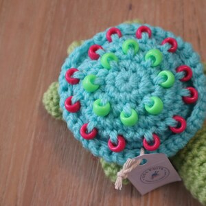 May include: A crocheted turtle toy with a turquoise shell adorned with red and green beads. The head and feet are green. A small tag is attached to the turtle. A handmade item.