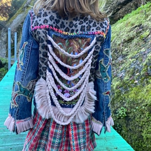 Può includere: Una giacca di jeans blu con un pannello in tessuto stampato leopardato sul retro. La giacca ha un dettaglio di corda intrecciata bianca e un bordo di volant rosa. La giacca è indossata sopra una gonna a quadri.