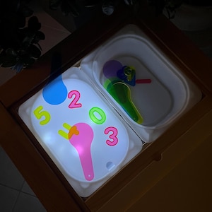 Peut inclure: Une table en bois avec deux plateaux en plastique blanc. Un plateau contient les chiffres éclairés 2, 3 et 5, et une forme rose. L'autre plateau contient des outils en plastique colorés. La scène est éclairée de l'intérieur des plateaux.