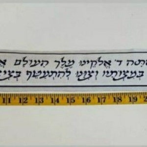 May include: A white fabric banner with blue Hebrew text and decorative designs. The text reads "Blessed are You, Lord our God, King of the Universe, who has sanctified us with His commandments and commanded us to wear tzitzit."