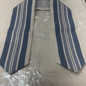 May include: A blue and white striped prayer shawl, or tallit, with a white center panel. The blue stripes are a medium shade and alternate with white stripes. The tallit is folded and laid on a white surface.