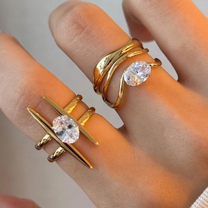 May include: Two gold rings are shown on a hand. One ring has a crisscross design with a clear gemstone. The other ring has a layered, wavy design, also with a clear gemstone. The rings are set against a neutral background.