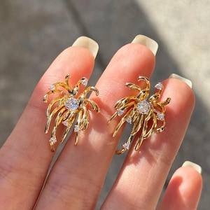 Puede incluir: Un par de pendientes dorados. Cada pendiente presenta una piedra preciosa central transparente rodeada de delicadas líneas doradas radiantes, con piedras preciosas transparentes más pequeñas que acentúan el diseño. Los pendientes se muestran en una mano.