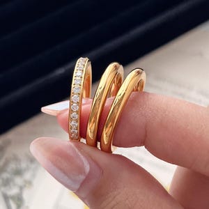 Könnte beinhalten: Drei goldene Ringe werden von einer Person gehalten. Ein Ring ist mit kleinen, klaren Steinen besetzt. Die anderen beiden Ringe sind glatt und glänzend. Die Ringe werden vor einem schwarzen Samthintergrund und einem Blumendruck präsentiert.