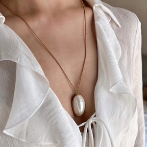 Puede incluir: Un collar dorado con un gran colgante ovalado de perla. El colgante está engastado en un marco dorado y cuelga de una fina cadena dorada. El collar se lleva sobre una blusa blanca con volantes.