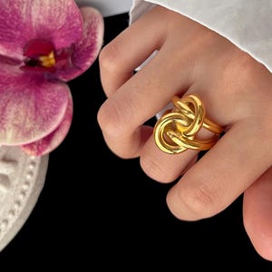 Peut inclure: Une bague dorée avec un motif de nœud. La bague est portée au doigt, avec une manche de chemise blanche en arrière-plan. Une fleur d'orchidée rose et violette est visible sur le côté gauche de l'image.