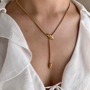 Könnte beinhalten: Goldfarbene Halskette mit schlangenähnlichem Design. Der Y-förmige Anhänger hat einen kleinen, klaren Stein und einen herabhängenden, spitz zulaufenden Anhänger. Die Halskette wird über einer weißen Bluse getragen.