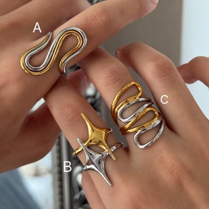 May include: Three rings are shown on fingers. Ring A is a gold and silver swirl design. Ring B is a gold and silver starburst design. Ring C is a gold and silver swirl design. The rings are made of metal.
