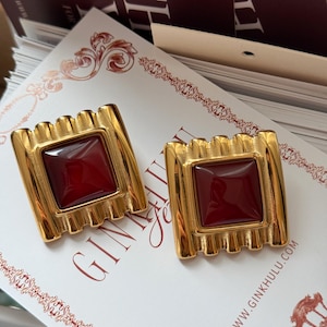 Könnte beinhalten: Ein Paar goldfarbene quadratische Ohrringe mit einem rechteckigen, tiefroten Mittelstein. Die Ohrringe haben ein geriffeltes Design an den Seiten und werden auf einer Karte mit dem Text "GINKHULU" und einer Website-Adresse präsentiert.