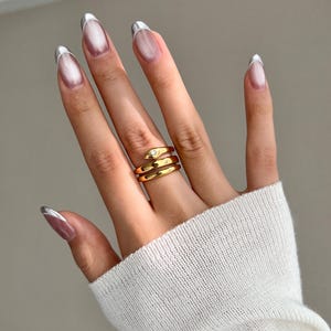 May include: A gold snake ring with a single clear stone on the head. The ring is worn on the middle finger of a hand with silver tipped nails.