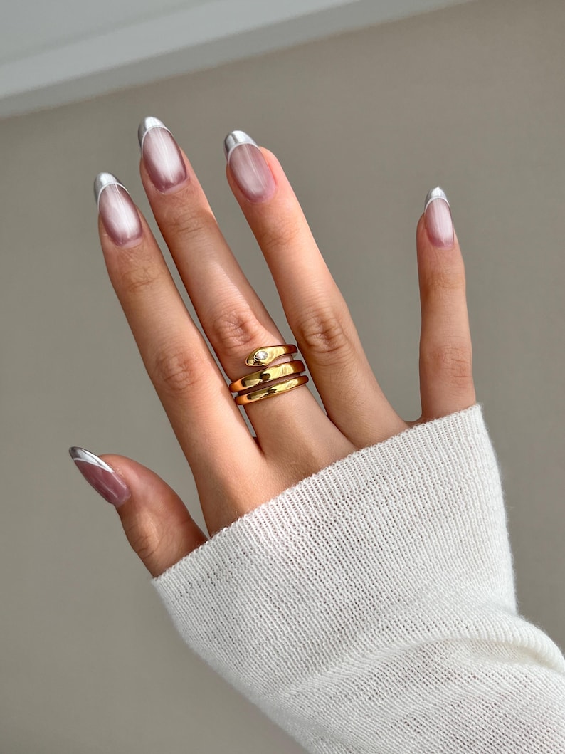 May include: A gold snake ring with a single clear stone on the head. The ring is worn on the middle finger of a hand with silver tipped nails.