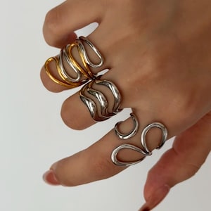May include: A hand displaying four abstract rings. One ring is gold and silver, the others are silver. The rings feature a fluid, organic design with curved lines and open spaces. The jewellery has a modern, minimalist aesthetic.