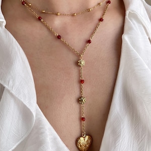 May include: A layered gold-toned necklace with red beads and a heart-shaped pendant. The necklace features a delicate chain with small gold beads and red beads. The pendant has an ornate floral design.