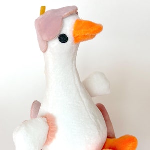 May include: A white plush goose toy with a pink hat and orange feet. The goose has a black eye and a small orange beak.