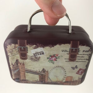 May include: A brown vintage-style suitcase with a silver handle and metal clasps. The suitcase has a printed design featuring the Eiffel Tower, a Ferris wheel, and the text "Ville de Paris".