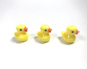 May include: Three small, bright yellow rubber ducks arranged in a horizontal row. Each duck features a smooth, glossy finish, a vibrant orange beak, and a single black dot for an eye. The figures are molded in a classic duck shape with a rounded body.