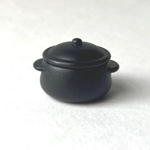 May include: A small, black, round pot with a lid and two side handles. The pot is made of a matte material and sits on a white surface. The lid has a small knob on top.
