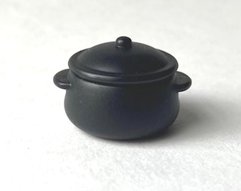 Op de afbeelding: Een kleine, zwarte pot met een deksel en twee handvatten aan de zijkant. De pot is rond en gemaakt van mat materiaal. Hij staat op een witte ondergrond. Het deksel heeft een kleine knop.