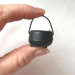 May include: A miniature black cauldron with a curved handle. The small pot has three tiny feet and is held between two fingers. The cauldron is a dark matte black and appears to be made of plastic.