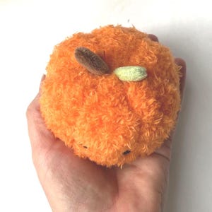 May include: A plush orange fruit toy with a fuzzy texture, brown stem, and a small green accent. The toy is held in a hand against a white background. The toy is a round shape.