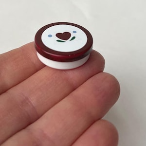 Puede incluir: Pequeño objeto decorativo redondo, con base blanca y parte superior roja oscura. La parte superior presenta un círculo blanco con un corazón marrón, puntos azules y hojas verdes. El objeto se sostiene en una mano.