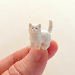 以下が含まれることがあります： 青い目をした白いプラスチック製の猫の小さな置物。猫は4本の足で立っており、尻尾は後ろに巻かれている。