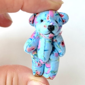 May include: A miniature teddy bear with a light blue floral pattern. The bear has a black nose, small ears, and a pink ribbon around its neck. The bear is held between two fingers.