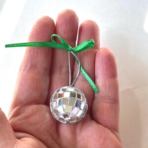 May include: A small, silver disco ball ornament with a green ribbon bow and silver string for hanging. The ball is covered in small, reflective squares. The ornament is held in a person's hand.