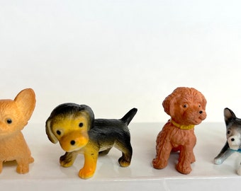 Könnte beinhalten: Vier kleine Plastikspielzeug-Hunde in verschiedenen Rassen und Farben. Die Hunde sind braun, schwarz, grau und orange.