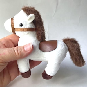 Puede incluir: Un juguete de peluche con forma de caballo blanco, con crin, cola y cascos marrones. Cuenta con una silla de montar y brida marrones. Un juguete suave para niños.
