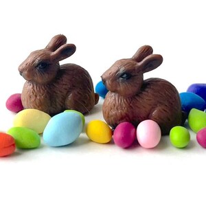 May include: Two brown rabbit figurines with detailed fur sit among colorful, pastel-colored Easter eggs. The eggs are in shades of pink, yellow, blue, green, and orange. The scene is set against a white background.