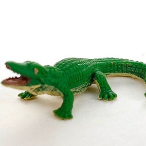 May include: A small, green plastic alligator toy with an open mouth, revealing a pink interior. The alligator has a textured, scaly body and is posed on a white surface. This toy is a miniature representation of a reptile.