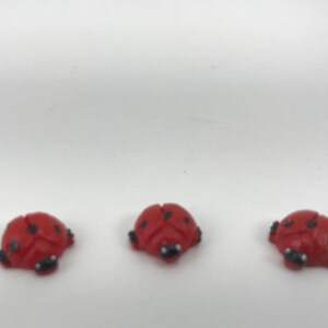 May include: Three small, red ladybug-shaped buttons or charms arranged in a horizontal row. Each ladybug features a smooth, rounded red body with distinct black spots and a small black head detail, creating a uniform set of three insect figures.