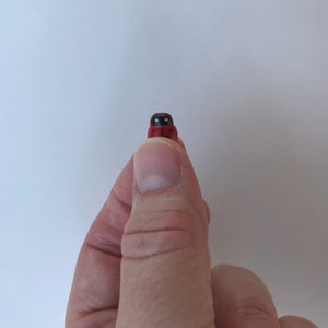May include: A tiny, miniature ladybug figurine featuring a bright red body with distinct black spots and a black head section with two small white markings, presented as a small decorative object held between a thumb and forefinger.