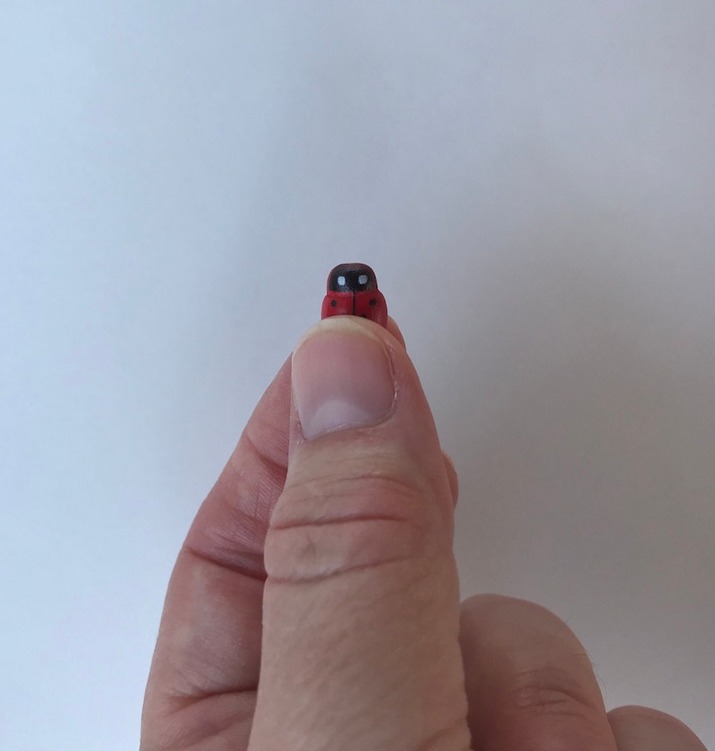 May include: A tiny, miniature ladybug figurine featuring a bright red body with distinct black spots and a black head section with two small white markings, presented as a small decorative object held between a thumb and forefinger.