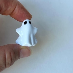 May include: A small, white ghost figurine with black eyes. The ghost has a simple, rounded shape and is held between two fingers. The figurine is likely ceramic or a similar material, and is a Halloween or spooky-themed item.