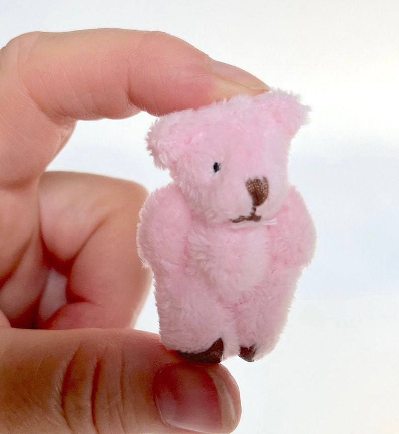 May include: A small pink plush teddy bear with brown eyes and a brown nose. The bear is being held by a hand.