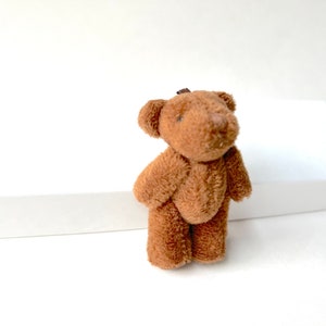 May include: A small brown teddy bear with a soft, plush texture. The bear is standing on a white surface.